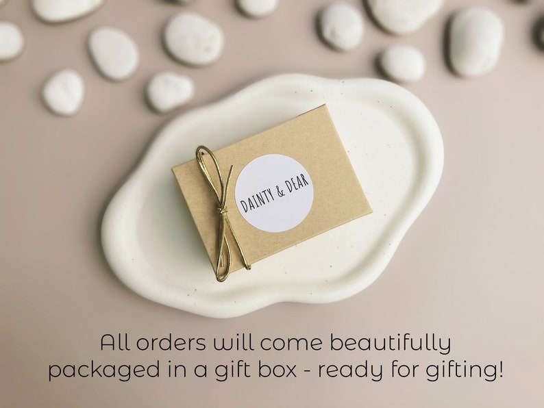 May include: A small brown box with a white circle sticker that says "DAINTY & DEAR" on it. The box is tied with a gold ribbon and is sitting on a white ceramic dish.