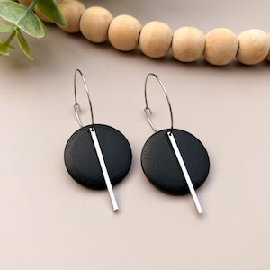 May include: A pair of black disc earrings with a silver bar accent. The earrings hang from silver hoops. The jewelry is displayed on a neutral background with wooden beads and greenery in the background. The earrings are a modern, minimalist design.