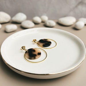 May include: A pair of gold hoop earrings with a brown and white tortoiseshell-patterned disc pendant hanging from each hoop.