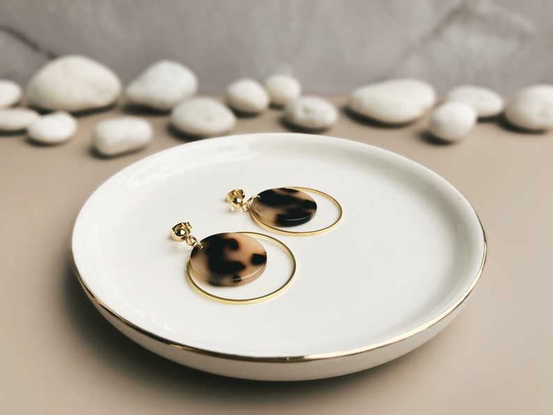 May include: A pair of gold hoop earrings with a brown and white tortoiseshell-patterned disc pendant hanging from each hoop.