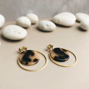 May include: A pair of gold hoop earrings with a brown and white tortoiseshell design. The earrings are on a light brown surface.