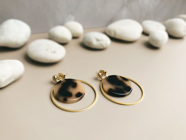 May include: A pair of gold hoop earrings with a brown and white tortoiseshell design. The earrings are on a light brown surface.