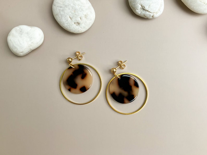 May include: A pair of gold hoop earrings with a brown and white patterned circle pendant hanging from each hoop.