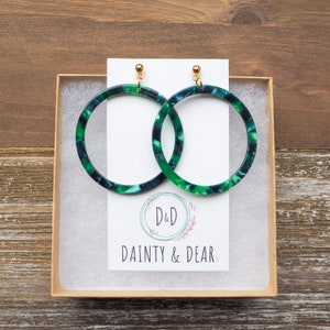 May include: A pair of large green and blue hoop earrings with a gold post. The earrings are in a box with a white card that says "D&D Dainty & Dear".