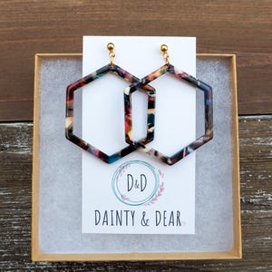 May include: A pair of hexagonal hoop earrings with a multi-colored marbled pattern. The earrings are made of acrylic and have gold-toned hooks. The earrings are packaged in a small white box with the text "D&D Dainty & Dear" printed on the front.