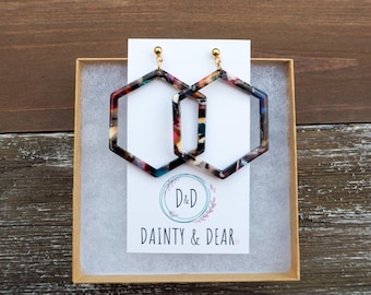 May include: A pair of hexagonal hoop earrings with a multi-colored marbled pattern. The earrings are made of acrylic and have gold-toned hooks. The earrings are packaged in a small white box with the text "D&D Dainty & Dear" printed on the front.