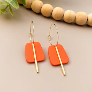 May include: A pair of orange and gold earrings. Each earring features a gold hoop and a rectangular orange pendant with a vertical gold bar. The earrings are displayed on a neutral background.
