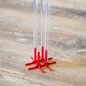 Puede incluir: Un soporte para tubos de ensayo de vidrio transparente con tres tubos. El soporte es rojo y tiene una base en forma de estrella. Los tubos son altos y cilíndricos, sobre una superficie de madera.