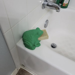 May include: A green frog-shaped soap holder holding a bar of soap. The soap holder is made of a textured material and is placed on the edge of a white bathtub. The frog has a detailed design.