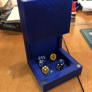 Peut inclure: Une tour à dés bleue de forme rectangulaire, avec une ouverture supérieure et un plateau inférieur. Plusieurs dés de différentes couleurs, dont noir, jaune et bleu, sont visibles à l'intérieur du plateau. La tour est conçue pour les jeux de société.