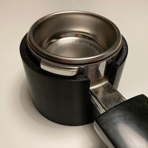 May include: A black espresso machine portafilter with a stainless steel filter basket. The portafilter has a black handle and is designed to hold ground coffee for brewing espresso. The filter basket is visible inside the portafilter.