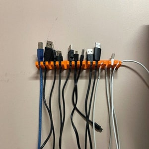 May include: An orange 3D-printed cable organizer mounted on a wall, holding various USB cables. The cables are black, white, and blue, with different connector types. The organizer has multiple slots to keep the cables neatly arranged.