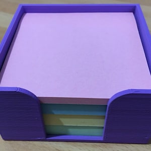 May include: A purple, square desk organizer holding a stack of pink sticky notes. The organizer has a curved cutout on the front for easy access to the notes. Below the pink notes are a few multi-colored sticky notes.