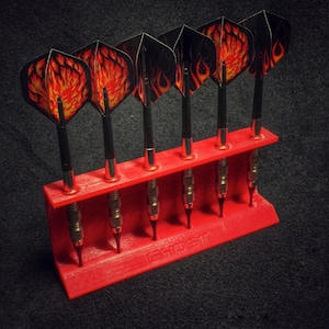 Może przedstawiać: Zestaw lotek z czarnymi trzonkami i lotkami z motywem płomieni, umieszczonych w czerwonym uchwycie. Uchwyt ma nadruk "GHOST". Lotki są starannie ułożone, gotowe do gry.