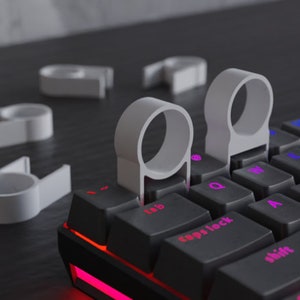May include: White keyboard key pullers are shown next to a black mechanical keyboard with illuminated keys. The key pullers are designed to remove and replace keyboard keys. The keyboard has a red underglow.