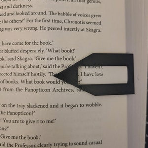 May include: A black, arrow-shaped bookmark made of plastic, resting on the pages of a book. The bookmark has a rectangular cutout in the center, and is used to mark a specific page in the book.