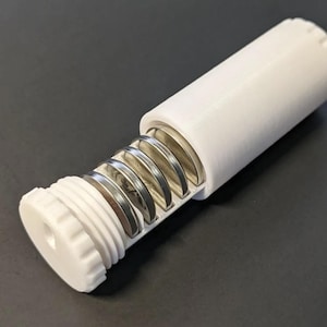 Puede incluir: Un contenedor cilíndrico blanco impreso en 3D con una tapa de rosca, parcialmente abierto para revelar una serie de discos metálicos plateados. El contenedor está sobre una superficie oscura. El diseño sugiere un mecanismo de almacenamiento o dispensación.