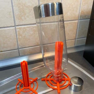 Puede incluir: Una botella de agua transparente con una tapa de acero inoxidable, un escurridor naranja y una tapa de acero inoxidable. La botella está dentro del escurridor naranja. Los artículos están sobre una superficie de acero inoxidable.