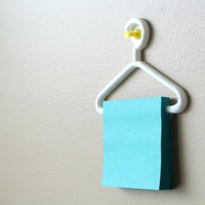 May include: A white, hanger-shaped wall-mounted memo holder with a yellow pin. A stack of blue sticky notes is held in place by the hanger. The memo holder is mounted on a beige wall.