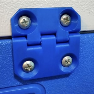 Può includere: Primo piano di una cerniera in plastica blu con quattro viti argentate. La cerniera è montata su una superficie bianca e blu. La cerniera è un componente utilizzato in mobili o altre applicazioni.