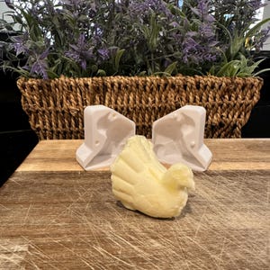 May include: A light yellow turkey-shaped soap sits on a wooden cutting board, with two white molds behind it. A woven basket with purple flowers is in the background. The soap has detailed feather and body features.