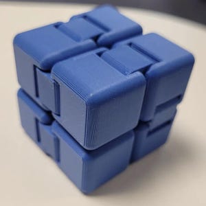 May include: A blue fidget cube toy. The cube is made of connected blocks that can be rotated and manipulated. The toy is a solid blue color and has a textured surface. The cube is designed to provide a tactile and engaging experience.