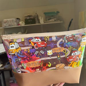 Puede incluir: Un estuche con cremallera que presenta un patr&oacute;n colorido e iridiscente de coches de carreras, se&ntilde;ales de tr&aacute;fico y paisajes des&eacute;rticos. El bolso tiene una base de piel sint&eacute;tica beige y un borde superior de tela a juego con acabado brillante.