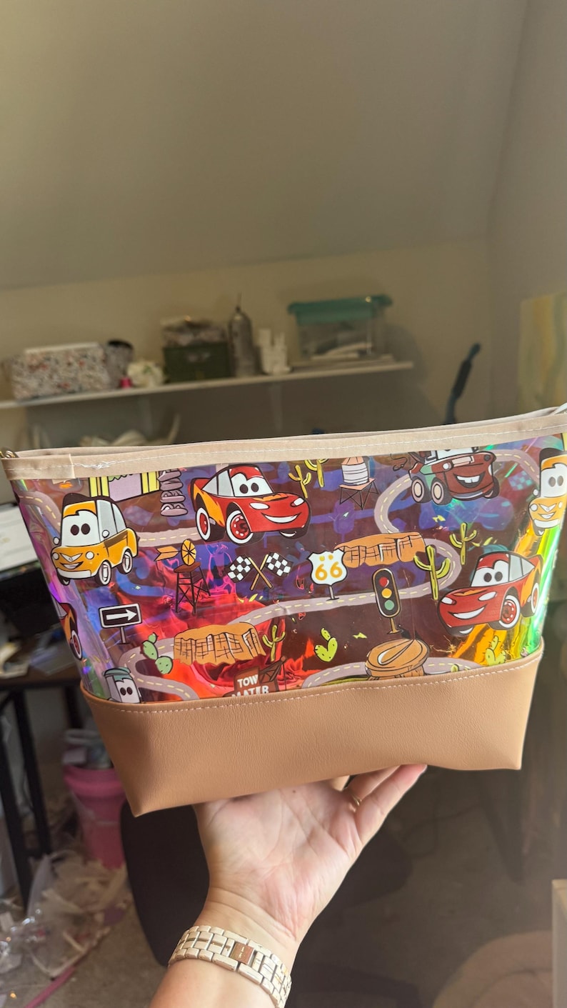 Puede incluir: Un estuche con cremallera que presenta un patr&oacute;n colorido e iridiscente de coches de carreras, se&ntilde;ales de tr&aacute;fico y paisajes des&eacute;rticos. El bolso tiene una base de piel sint&eacute;tica beige y un borde superior de tela a juego con acabado brillante.