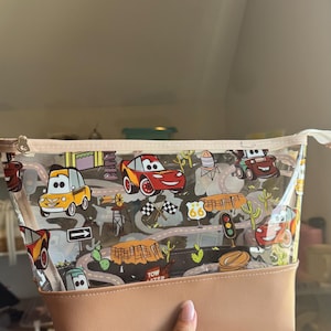Puede incluir: Un estuche transparente con cremallera que presenta un estampado de dibujos animados de coches de carreras, se&ntilde;ales de tr&aacute;fico, cactus y paisajes des&eacute;rticos. La bolsa tiene una base de piel sint&eacute;tica color canela y un borde a juego.
