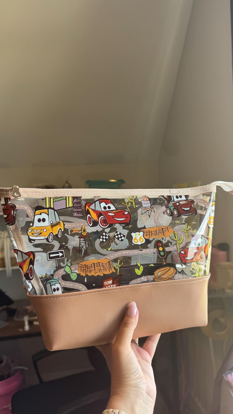 Puede incluir: Un estuche transparente con cremallera que presenta un estampado de dibujos animados de coches de carreras, se&ntilde;ales de tr&aacute;fico, cactus y paisajes des&eacute;rticos. La bolsa tiene una base de piel sint&eacute;tica color canela y un borde a juego.