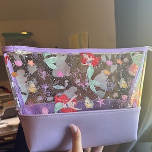 Puede incluir: Un neceser transparente y brillante con un estampado de la sirenita Ariel y el pez Flounder. El bolso tiene una base s&oacute;lida de color morado claro y un borde de cremallera a juego en la parte superior, ofreciendo un estuche de almacenamiento estructurado.