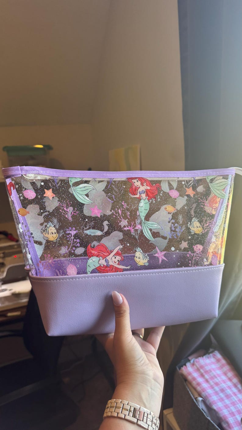 Puede incluir: Un neceser transparente y brillante con un estampado de la sirenita Ariel y el pez Flounder. El bolso tiene una base s&oacute;lida de color morado claro y un borde de cremallera a juego en la parte superior, ofreciendo un estuche de almacenamiento estructurado.
