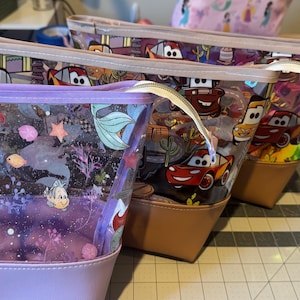 Puede incluir: Tres estuches con cremallera, partes superiores de vinilo transparente y bases de colores s&oacute;lidos. El estuche delantero es de color morado claro con un estampado de Ariel y Flounder. Los dos estuches traseros tienen un estampado de Cars con coches de carreras rojos y amarillos.