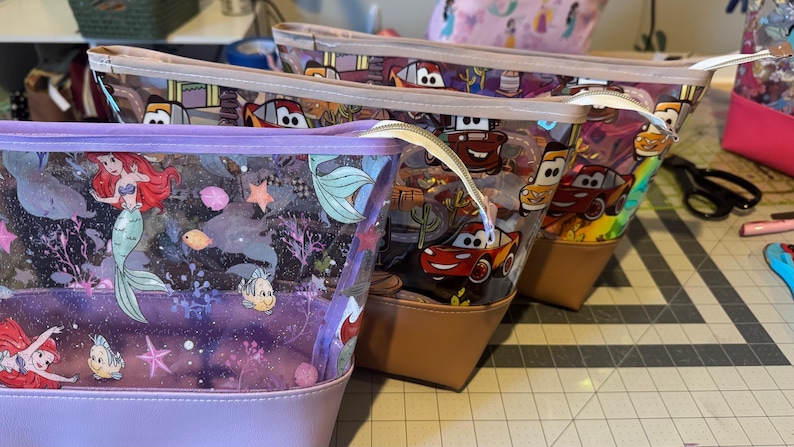 Puede incluir: Tres estuches con cremallera, partes superiores de vinilo transparente y bases de colores s&oacute;lidos. El estuche delantero es de color morado claro con un estampado de Ariel y Flounder. Los dos estuches traseros tienen un estampado de Cars con coches de carreras rojos y amarillos.
