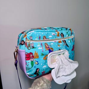 Könnte beinhalten: Eine hellblaue Wickeltasche mit Reißverschluss und einem Feuchttücher-Spender. Die Tasche hat einen farbenfrohen Aufdruck von Disney-Prinzessinnen und anderen Figuren. Sie hat eine lila Seitenwand und einen goldenen Reißverschluss.