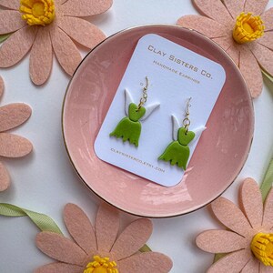 May include: A pair of green and white fairy-shaped earrings on a white card with the text "CLAY SISTERS CO. HANDMADE EARRINGS" and the website "CLAY SISTERCO.ETSY.COM".
