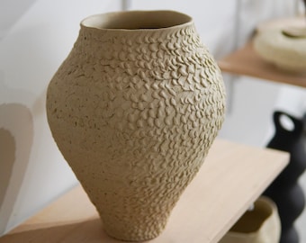 May include: A beige ceramic vase with a textured surface. The vase has a wide base and a narrow neck.