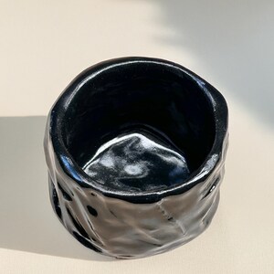 May include: A black ceramic planter with a textured, rough exterior. The planter has a wide, round opening and a smooth, glossy interior.