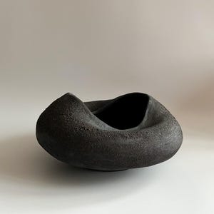 Puede incluir: Un cuenco de cerámica gris oscuro y texturizado con un diseño retorcido único. El cuenco tiene un acabado rugoso y mate y un interior profundo. La forma general es orgánica y escultural, adecuada para uso decorativo.