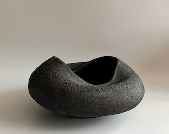 May include: A dark gray, textured ceramic bowl with a unique, twisted design. The bowl has a rough, matte finish and a deep interior. The overall shape is organic and sculptural, suitable for decorative use or as a centerpiece.