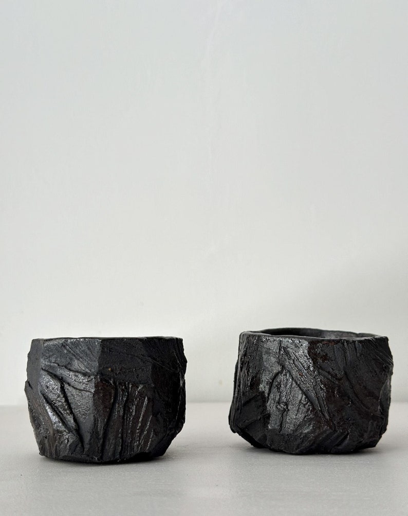 May include: Two black ceramic cups with a rough, textured surface. The cups are similar in shape and size, and they have a rustic, handmade look.