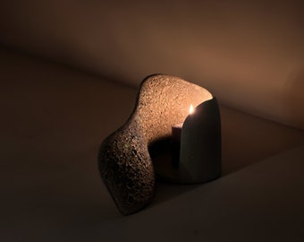 May include: A dark gray ceramic candle holder with a textured surface. The holder is shaped like a wave and has a small opening for a candle. A lit candle is inside the holder.