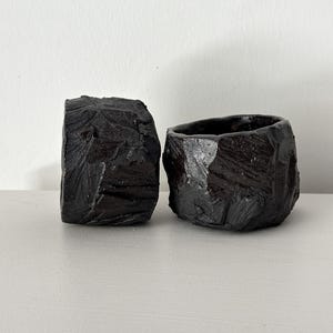 May include: Two small black ceramic planters with a rough, textured surface. The planters are made in a style that resembles rock formations.