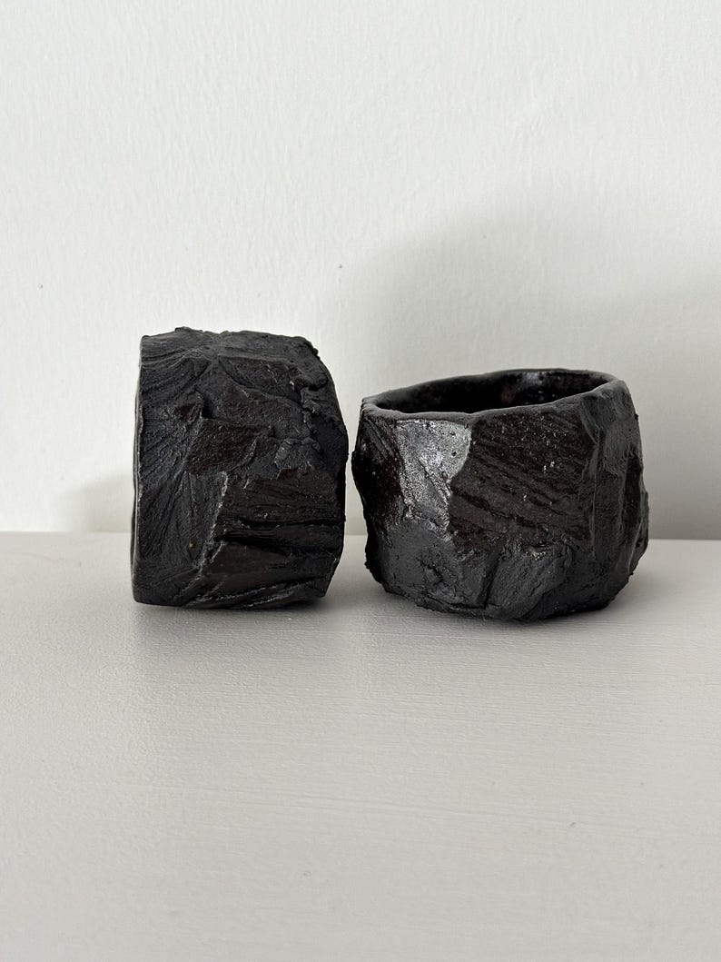 May include: Two small black ceramic planters with a rough, textured surface. The planters are made in a style that resembles rock formations.