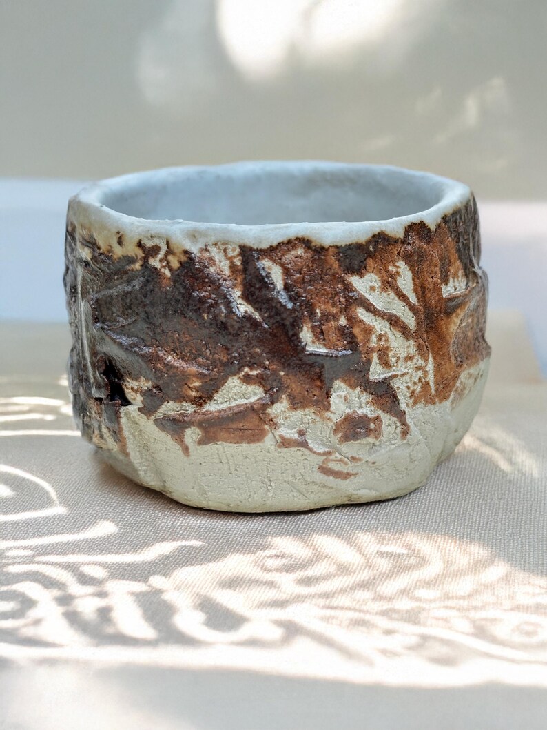 May include: A small, white ceramic bowl with a brown glaze. The glaze has a textured, rough appearance, giving the bowl a rustic look.