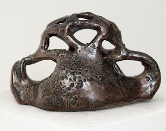 May include: A bronze-colored, abstract sculpture with an organic, porous design. The artwork features multiple openings and a textured surface, creating a unique and artistic aesthetic. The sculpture is displayed on a white surface.
