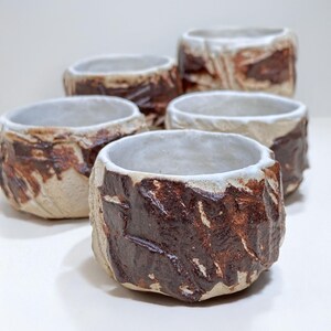 May include: Five small ceramic cups with a white glaze and brown accents. The cups have a rough, textured surface and are arranged in a row.