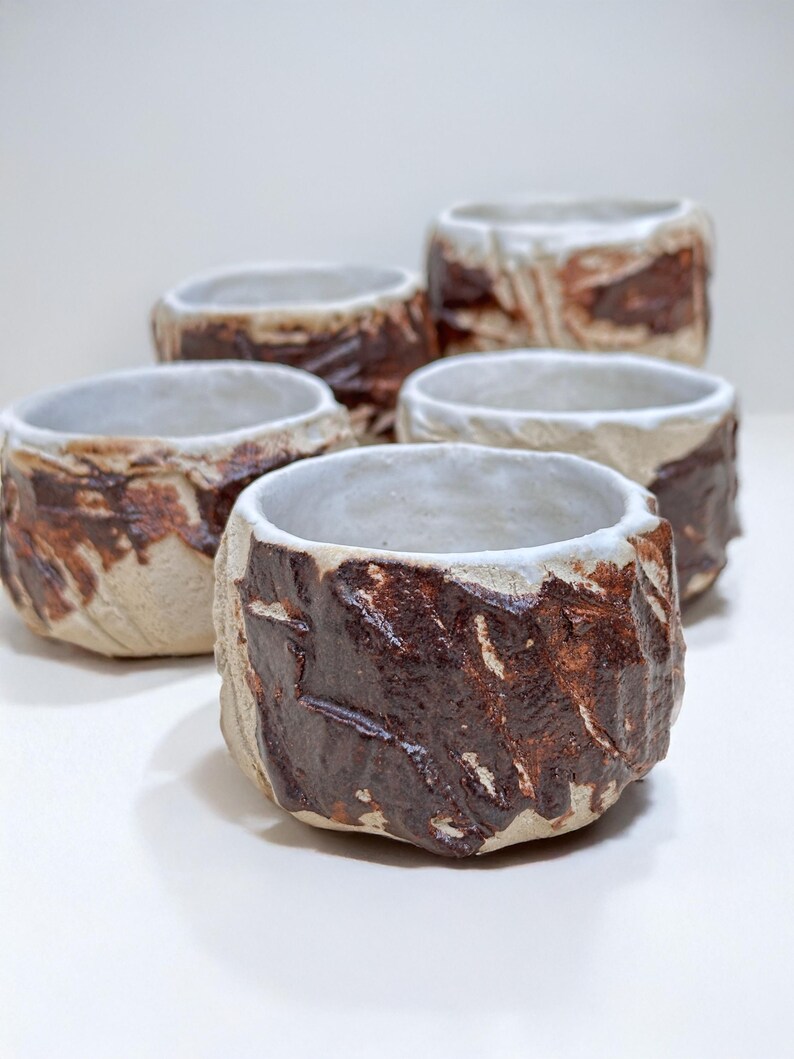 May include: Five small ceramic cups with a white glaze and brown accents. The cups have a rough, textured surface and are arranged in a row.