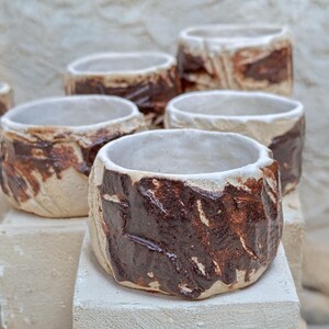 May include: Five ceramic cups with a white glaze and brown accents. The cups are arranged in a staggered pattern on a white surface.