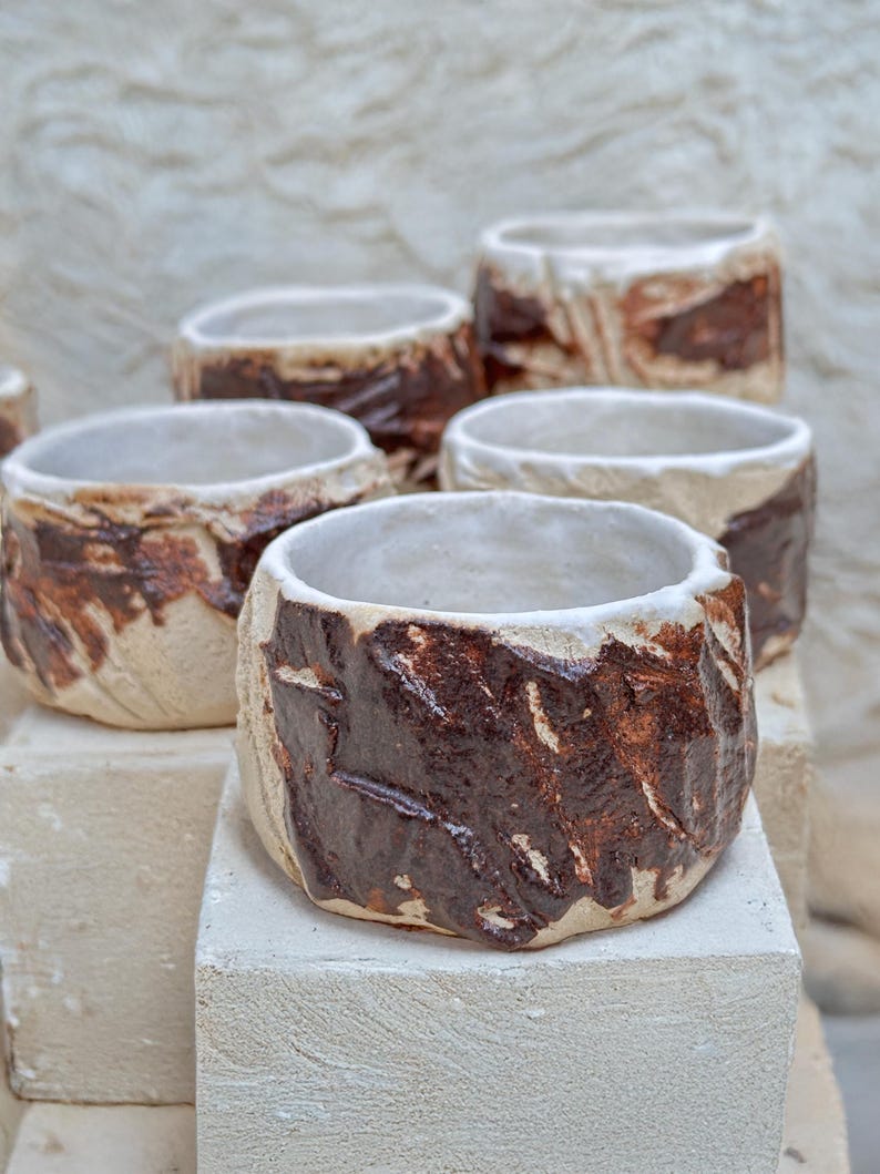 May include: Five ceramic cups with a white glaze and brown accents. The cups are arranged in a staggered pattern on a white surface.